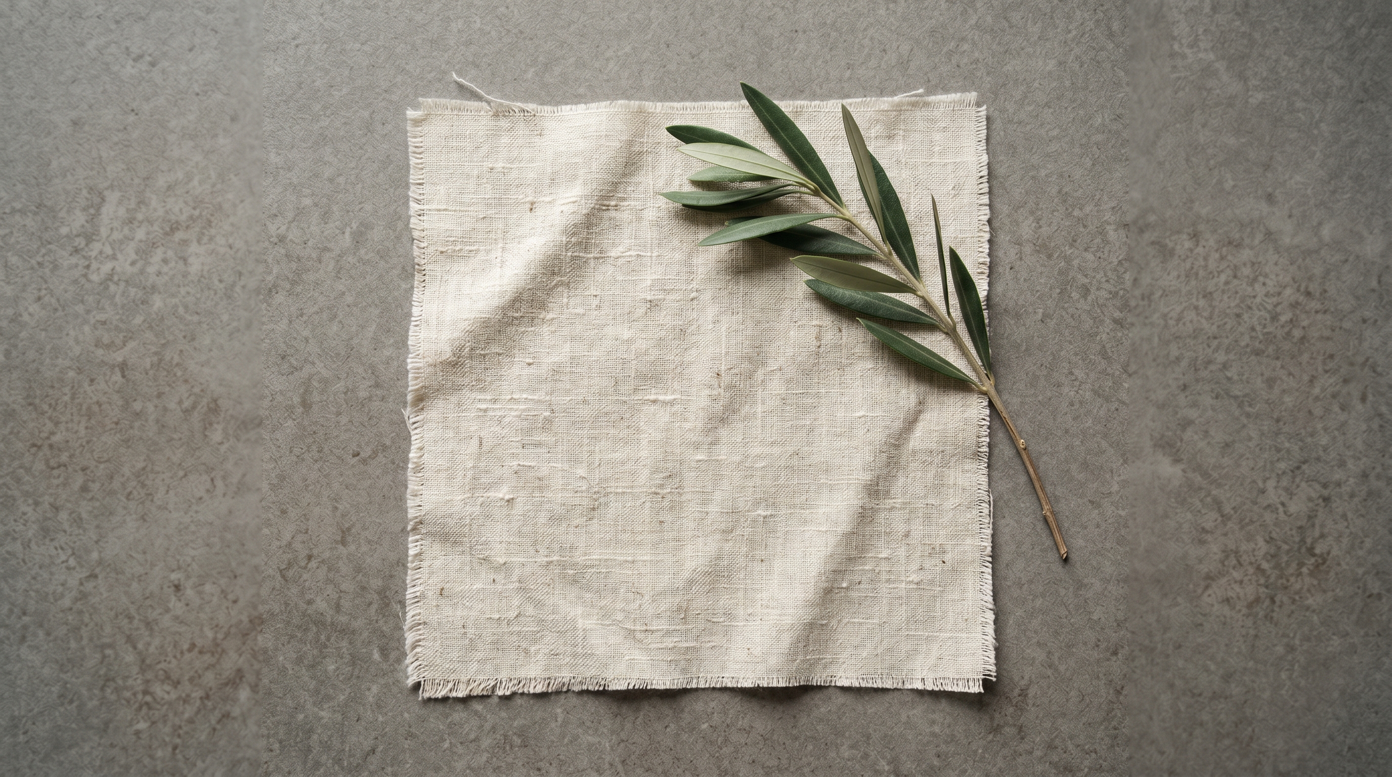 Warm bone-coloured raw-linen square showing natural slubs and weave irregularities in directional light, with an olive sprig, abstract texture study