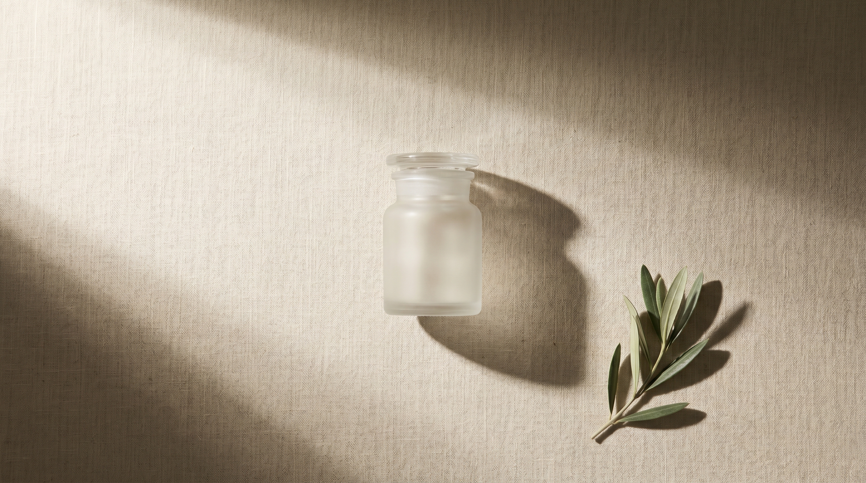 Single small frosted-glass apothecary jar precisely centred on warm bone-linen with a single olive sprig, visual metaphor for verified budget-tier selection