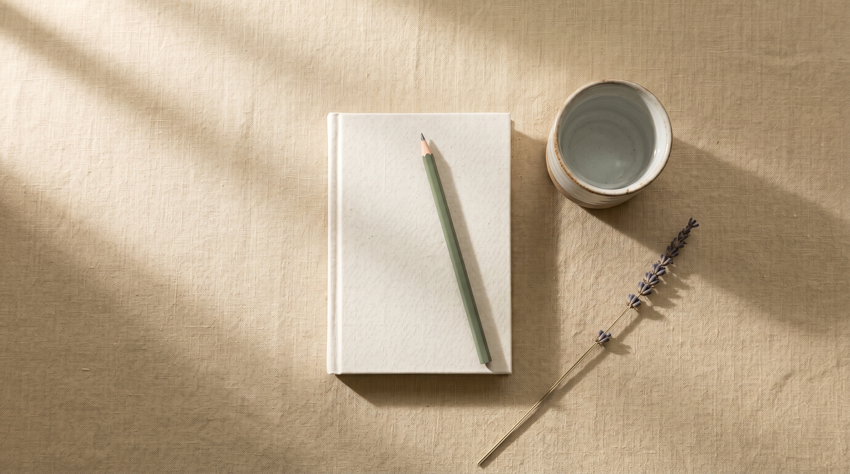 Bone-linen-bound notebook with a sage pencil, ceramic cup of water, and dried lavender on a warm linen surface, calm preparing-the-week-ahead mood
