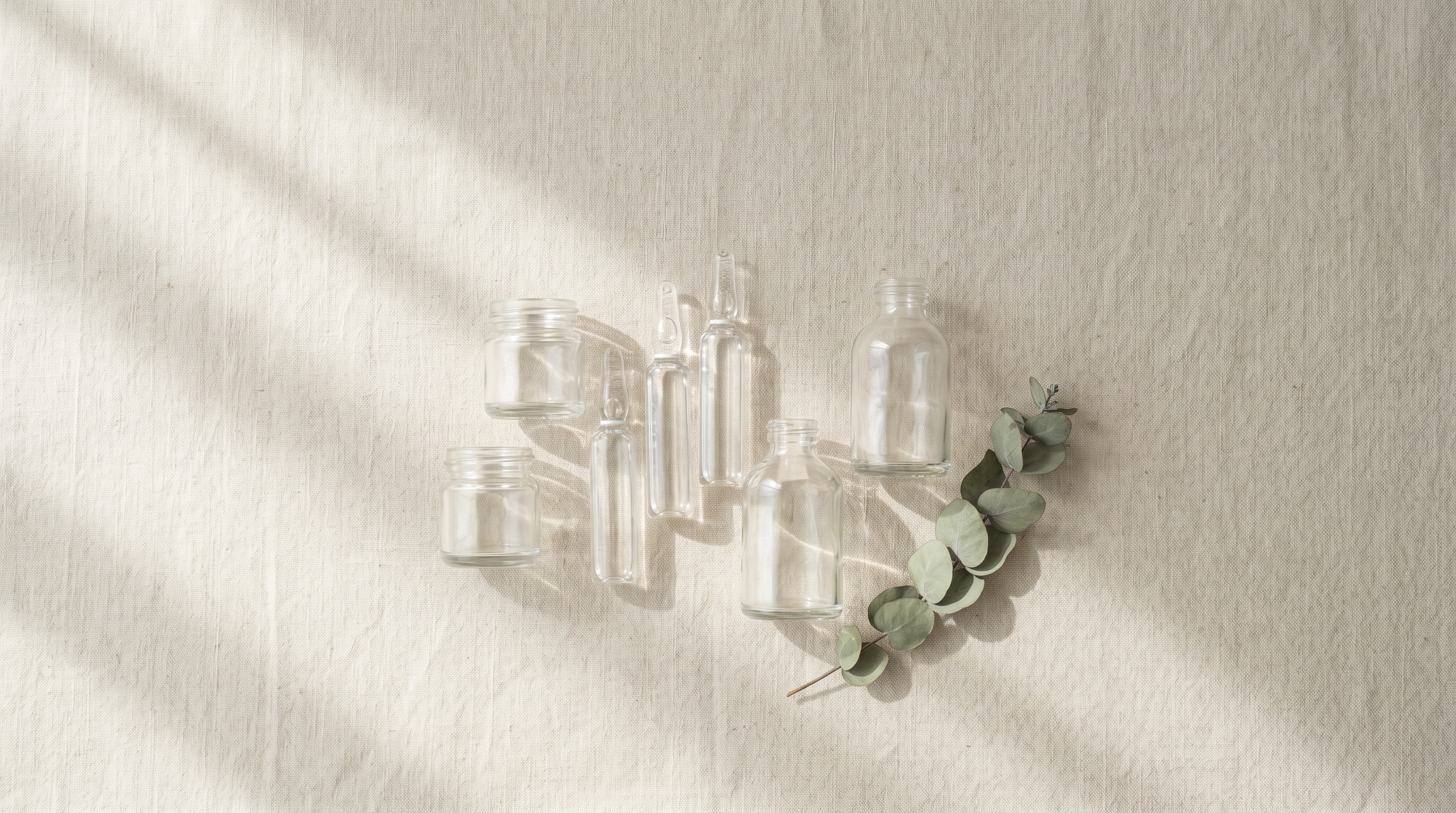 Seven small unlabelled glass vessels in varied shapes clustered on warm bone-coloured linen, visual taxonomy of the Korean skin booster menu